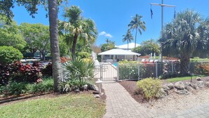 Un sentier mène à un espace piscine agrémenté d’un gazebo blanc, entouré de palmiers et de diverses plantes.