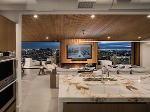 A modern living room with a large TV, comfortable seating, a kitchen area, and a city view.