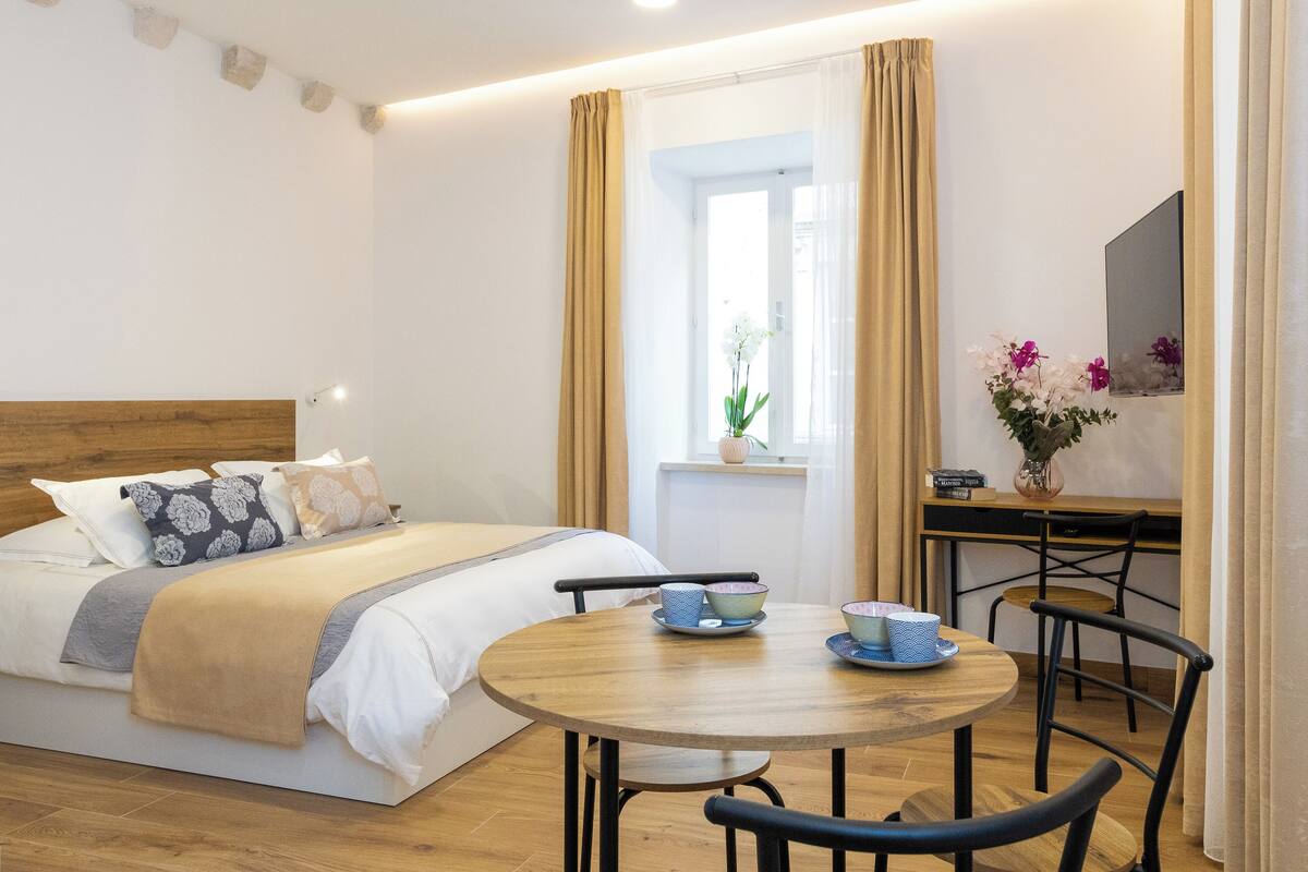 A bedroom with a bed, bedside table, and a small dining area with chairs and a table.