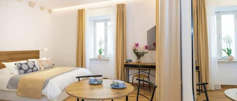 A modern hotel room with a large bed, a dining table with chairs, a desk with a chair, and a TV mounted on the wall.