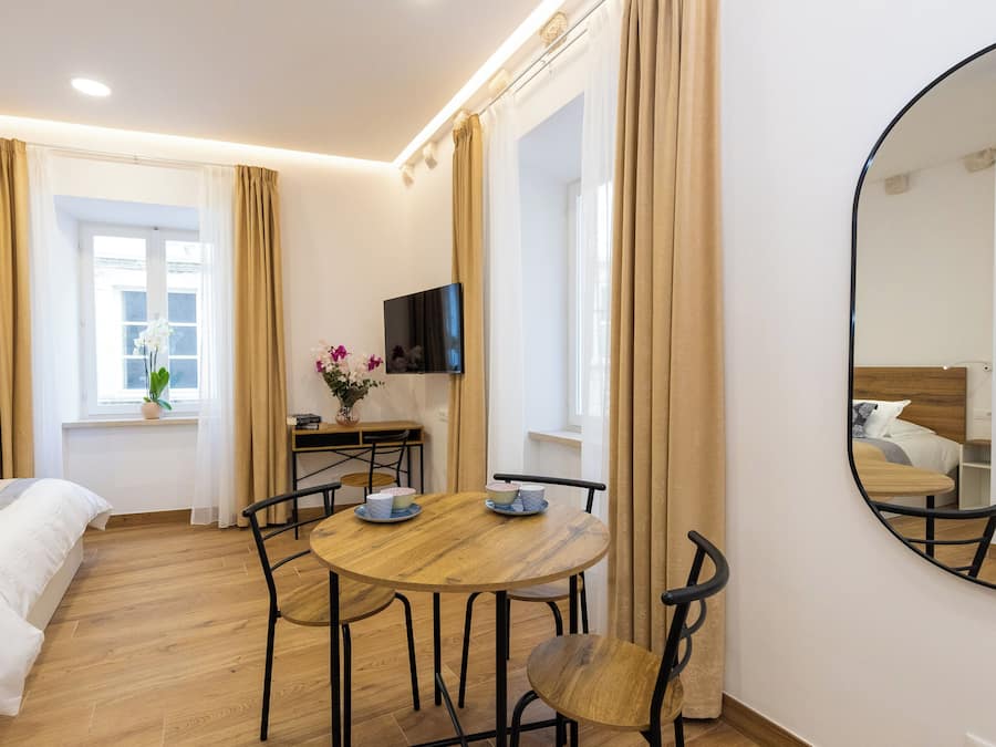 A modern hotel room with a wooden dining table, chairs, a TV, a bed, and a window with curtains.