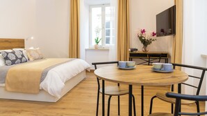 Une chambre d’hôtel avec un lit, une table ronde en bois entourée de chaises, une petite table sur laquelle est posé un vase de fleurs, et une fenêtre avec des rideaux.