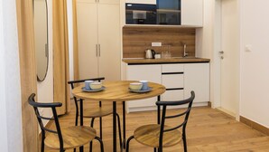 Une cuisine compacte avec une table ronde en bois et quatre chaises, un petit plan de travail avec une bouilloire, et un meuble suspendu au-dessus de l’évier.
