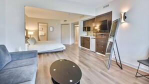 A modern hotel room with a bed, a sofa, a small round table, a kitchen area with a fridge, and a TV.
