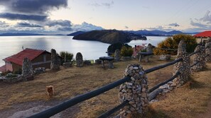 A rustic stone fence, a small stone building with a red roof, and a view of a lake with mountains in the background. - Kalluchi Ecolodge SunIsland TiticacaLake (Isla del Sol)