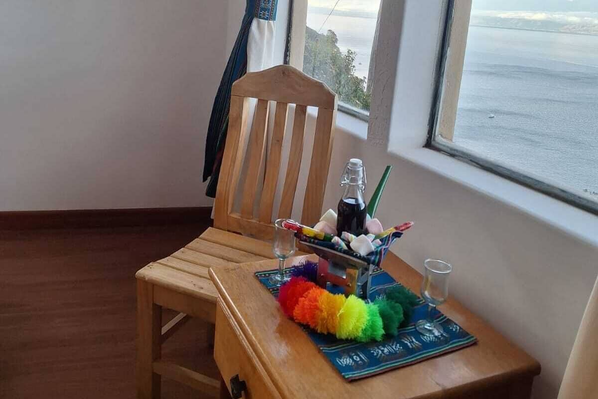A wooden chair and table with a bottle, glasses, and colorful items by a window with a view of the sea.