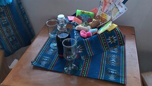 A table with a bottle, glasses, and a colorful mat. A window reveals a view of the sea and distant land. - Kalluchi Ecolodge SunIsland TiticacaLake (Isla del Sol)