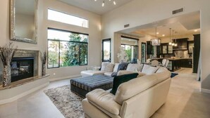 A spacious living room with a fireplace, large windows, and a modern kitchen in the background. - Luxury Contemporary Sanctuary w/  Pool and Views! (Fountain Hills)