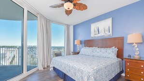 Un dormitorio con una cama grande, ventilador de techo, mesita de noche con lámparas, cómoda de madera y un cuadro en la pared.