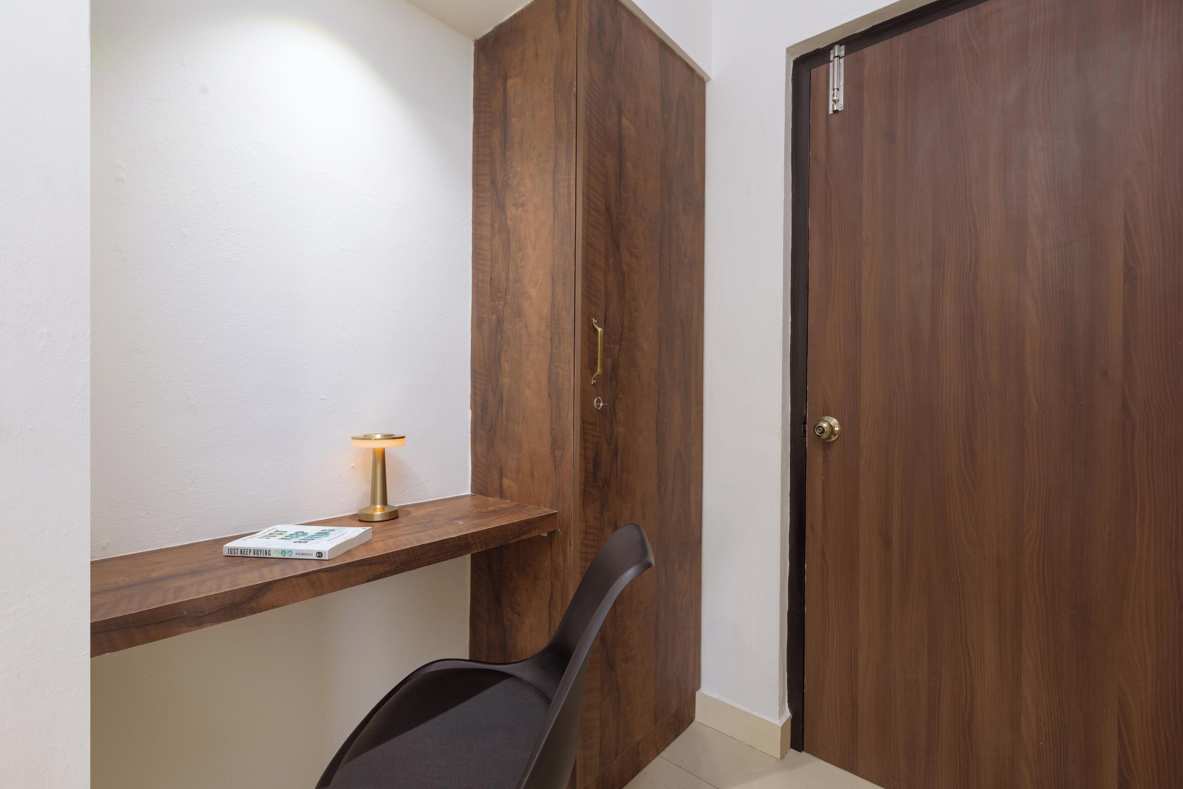 A room with a wooden wardrobe, a small wooden shelf with a book and a lamp, and a black chair.