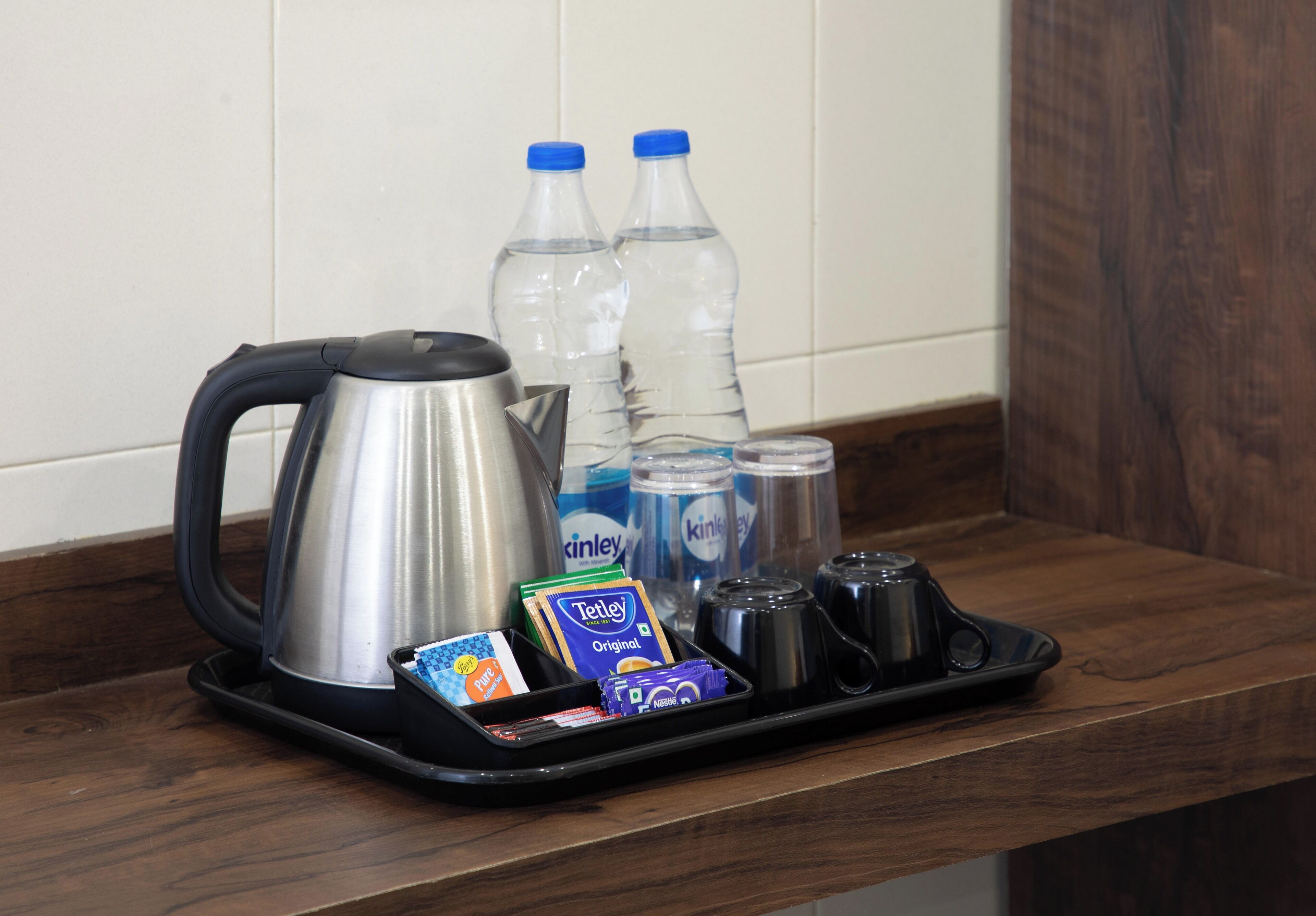 A tray with a kettle, bottled water, coffee cups, and tea packets on a wooden counter.