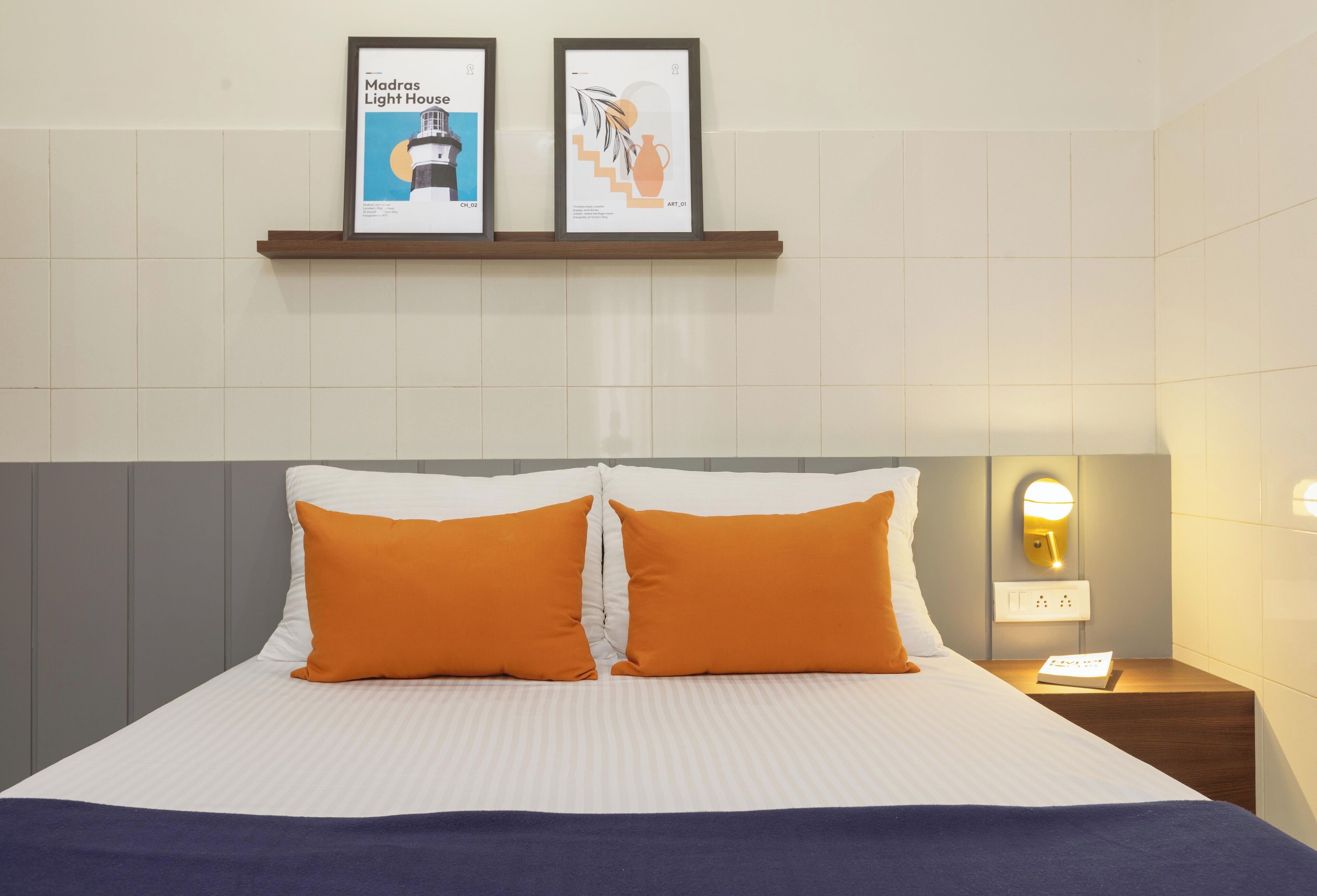 Una habitació d'hotel amb un llit, dos coixins taronja, una tauleta de nit amb una làmpada i dos quadres al mur.