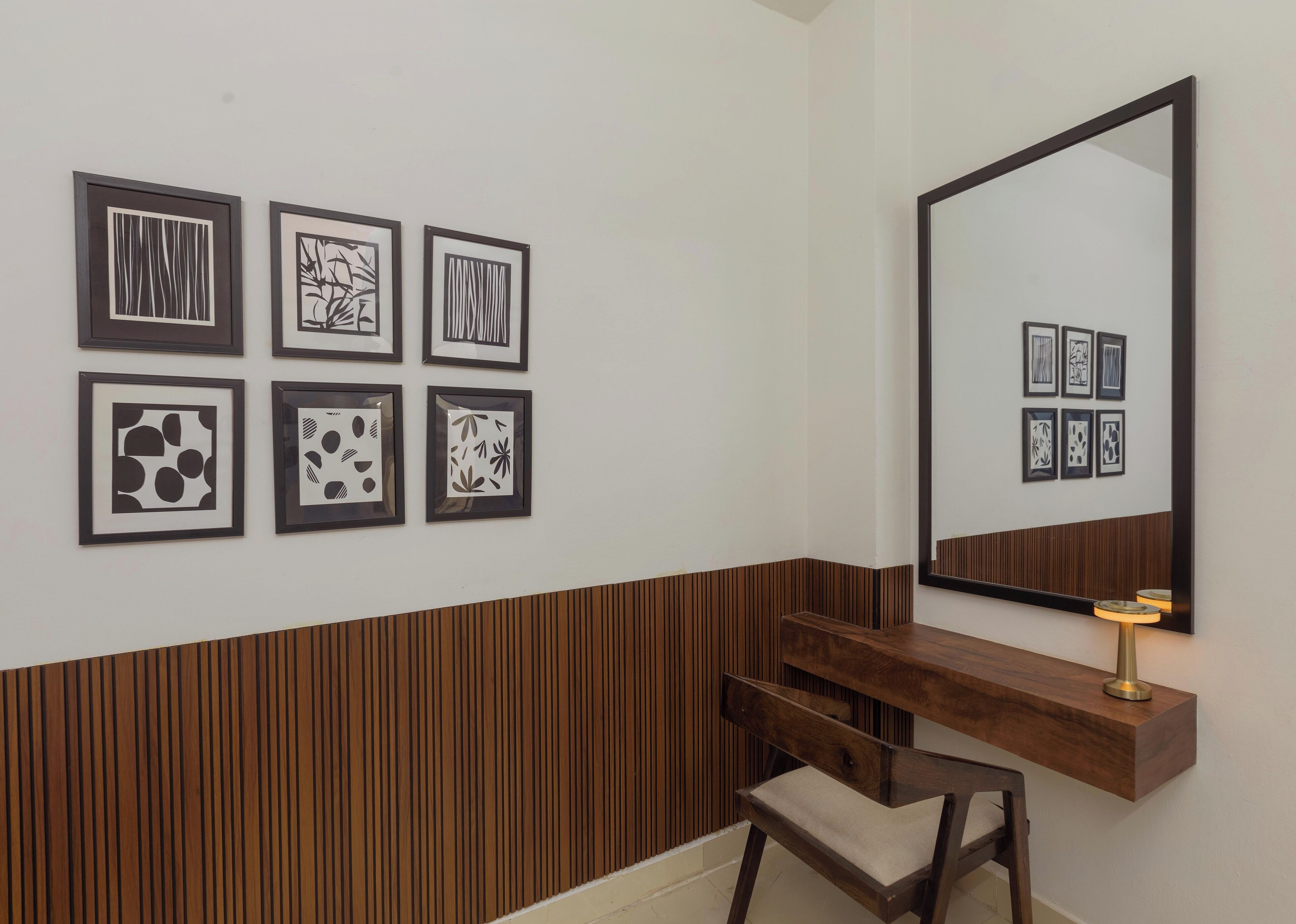 Una pared con seis obras de arte abstractas enmarcadas, un banco de madera, un espejo y una pequeña mesa con una lámpara.