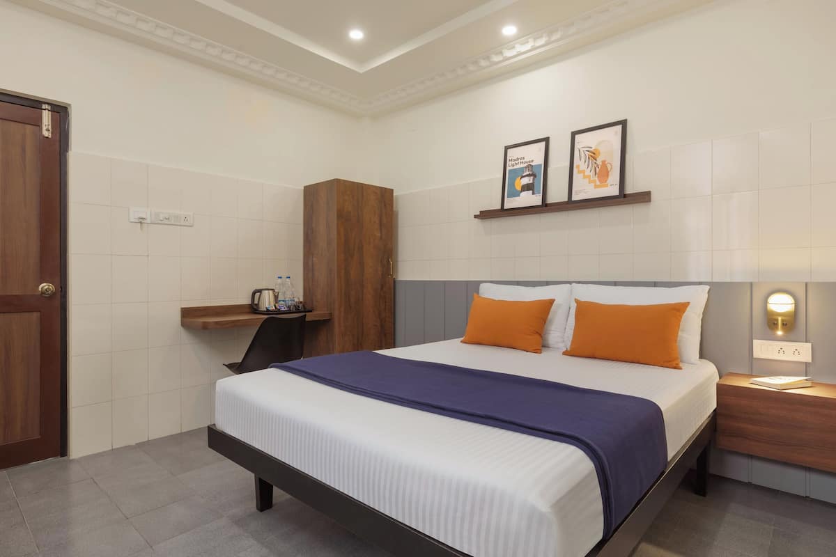 A hotel room with a bed, a wooden wardrobe, a nightstand with a lamp, and a shelf with a framed picture.