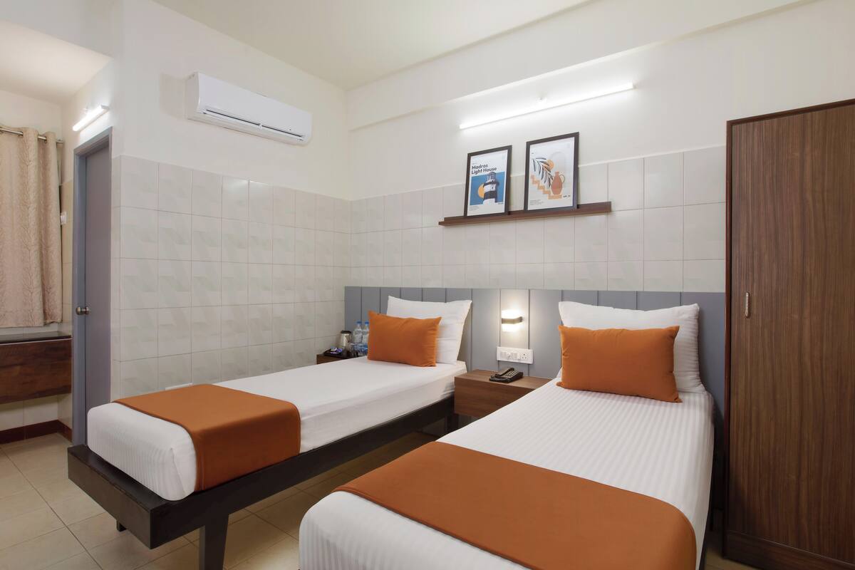 A hotel room with two beds, a wooden wardrobe, a bathroom with a shower, and a wall-mounted air conditioner.