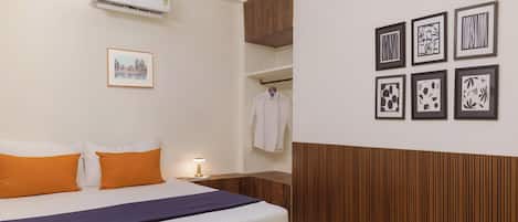 A hotel room with a bed, a ceiling fan, an air conditioner, a wall-mounted shelf with a white shirt, and framed artwork on the wall.