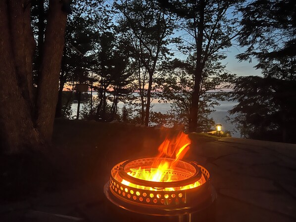 A campfire with a metal grill, surrounded by trees and a body of water in the background.