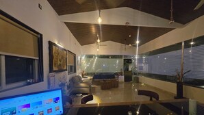 A modern living room with a large flat-screen TV, a sofa, a coffee table, and a dining area with a glass table and chairs. - SAAZ RESORT AND LAWN (Khalapur)