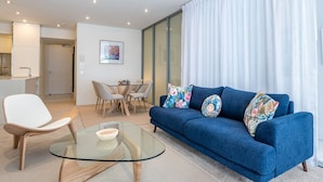 A modern living room with a blue sofa, a glass coffee table, and a white chair. There is a dining area with a table and chairs in the background. - WaterEdge West End Retreat (West End)