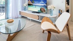 A modern living room with a glass table, a wooden chair, a flat-screen TV, and a view of a tropical beach. - WaterEdge West End Retreat (West End)