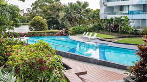 A swimming pool with a wooden deck, surrounded by lush greenery and palm trees. - Riverside at WaterEdge West End (West End)