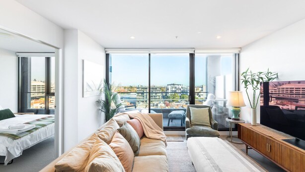 A modern living room with a sofa, a TV, and a view of the city. - Wellness and Style - 2BR Apartment with Pool, Hot Tub and Gym (Moonee Ponds)