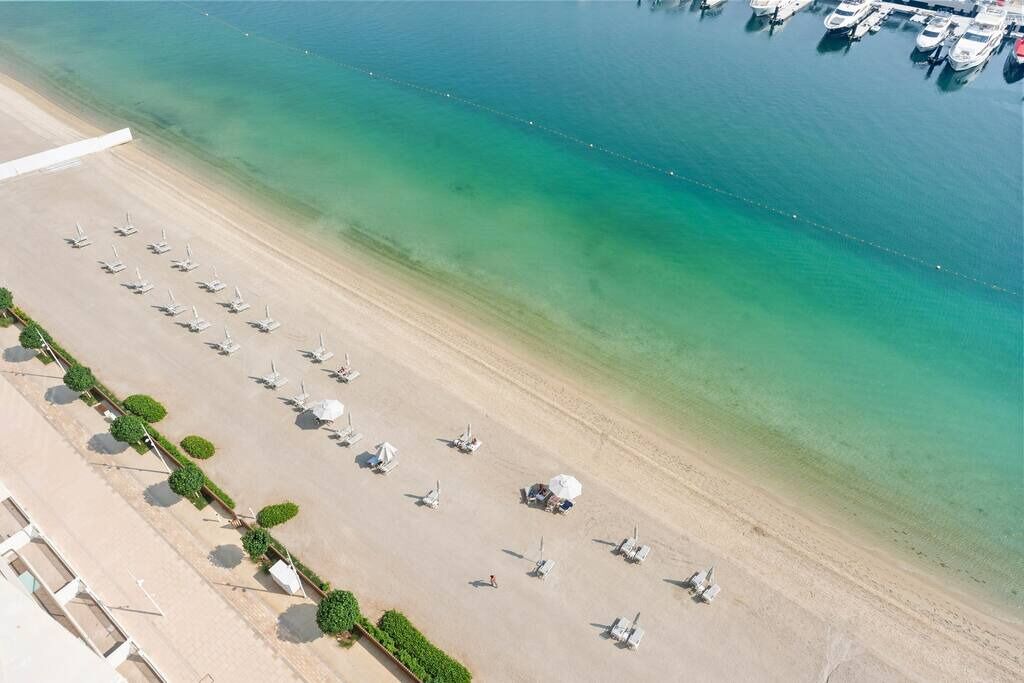 Vue aérienne d’une plage bordée de rangées de transats et d’un port de plaisance où sont amarrés des bateaux.
