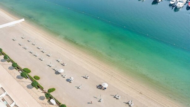 Vue aérienne d’une plage bordée de rangées de transats et d’un port de plaisance où sont amarrés des bateaux.
