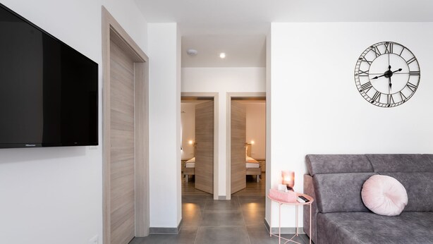 A modern living room with a grey sofa, a pink cushion, a wall-mounted clock, and a flat-screen TV on the wall. - Apartment Kurte (Nerezise)