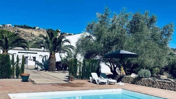 A swimming pool with clear blue water, surrounded by a patio area with lounge chairs and a stone wall. - Hotel Rural La Hacienda de Ronda  (Ronda)