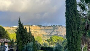 A courtyard with a stone fountain, a wooden fence, and a view of a hilly landscape with trees and a building in the distance. - Hotel Rural La Hacienda de Ronda  (Ronda)