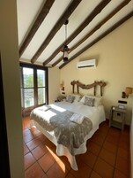 A bedroom with a large bed, wooden beams, an air conditioning unit, and a view through a glass door.