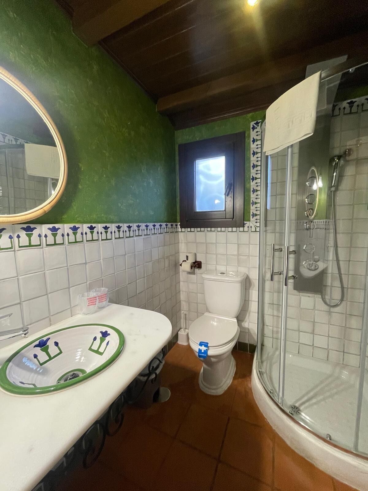 Un baño con pared verde, baldosas blancas, un espejo redondo, un lavabo con borde verde, un inodoro y una ducha con cerramiento de vidrio.