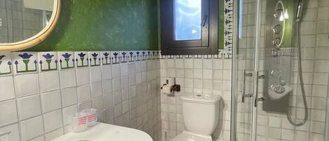 Une salle de bain avec un mur vert, des carreaux blancs, un miroir rond, un lavabo avec un bord vert, des toilettes et une douche fermée par une paroi de verre.