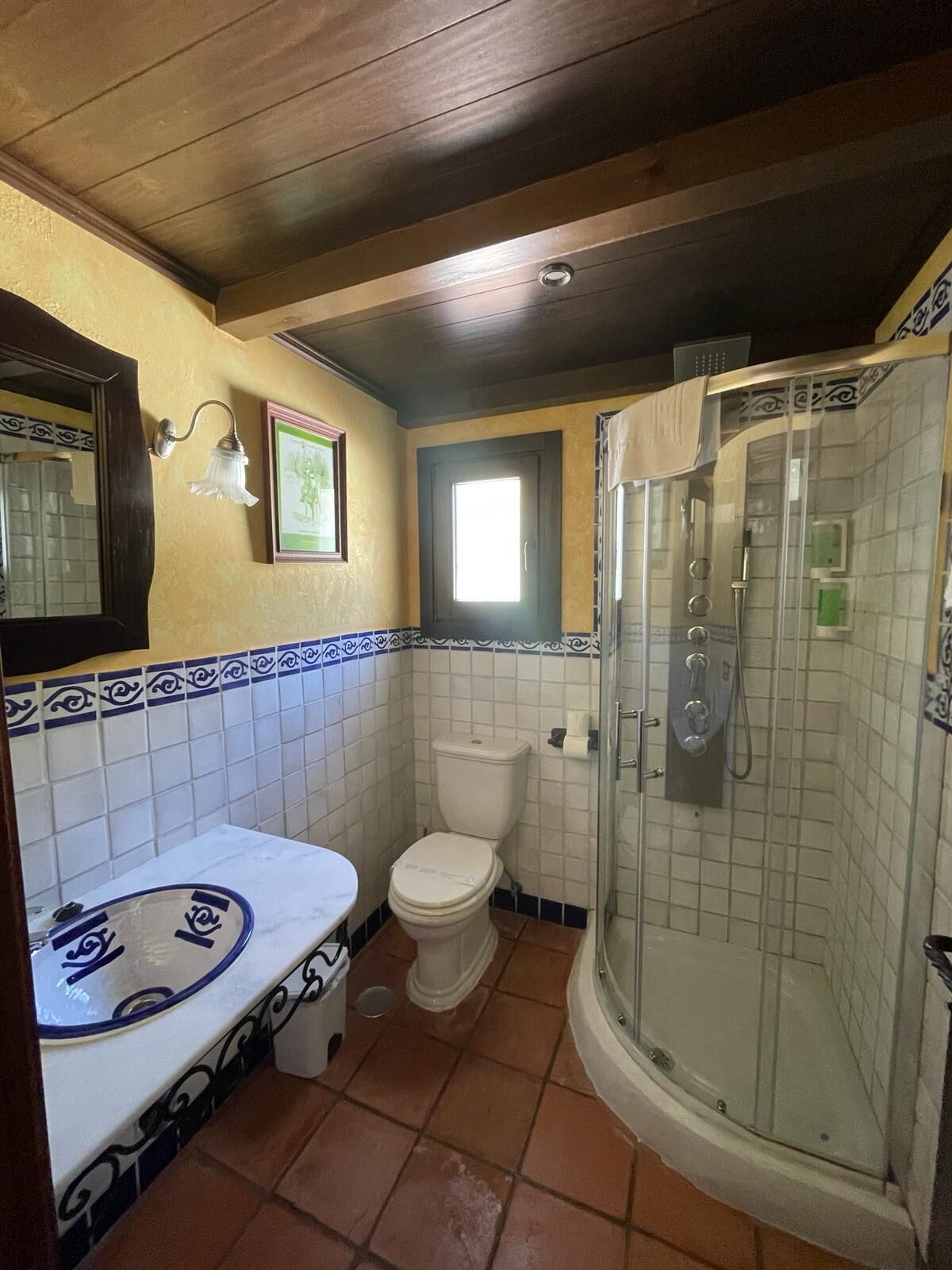 Un baño con lavamanos, inodoro y ducha blancos. Hay una ventana sobre el inodoro y un espejo sobre el lavamanos.