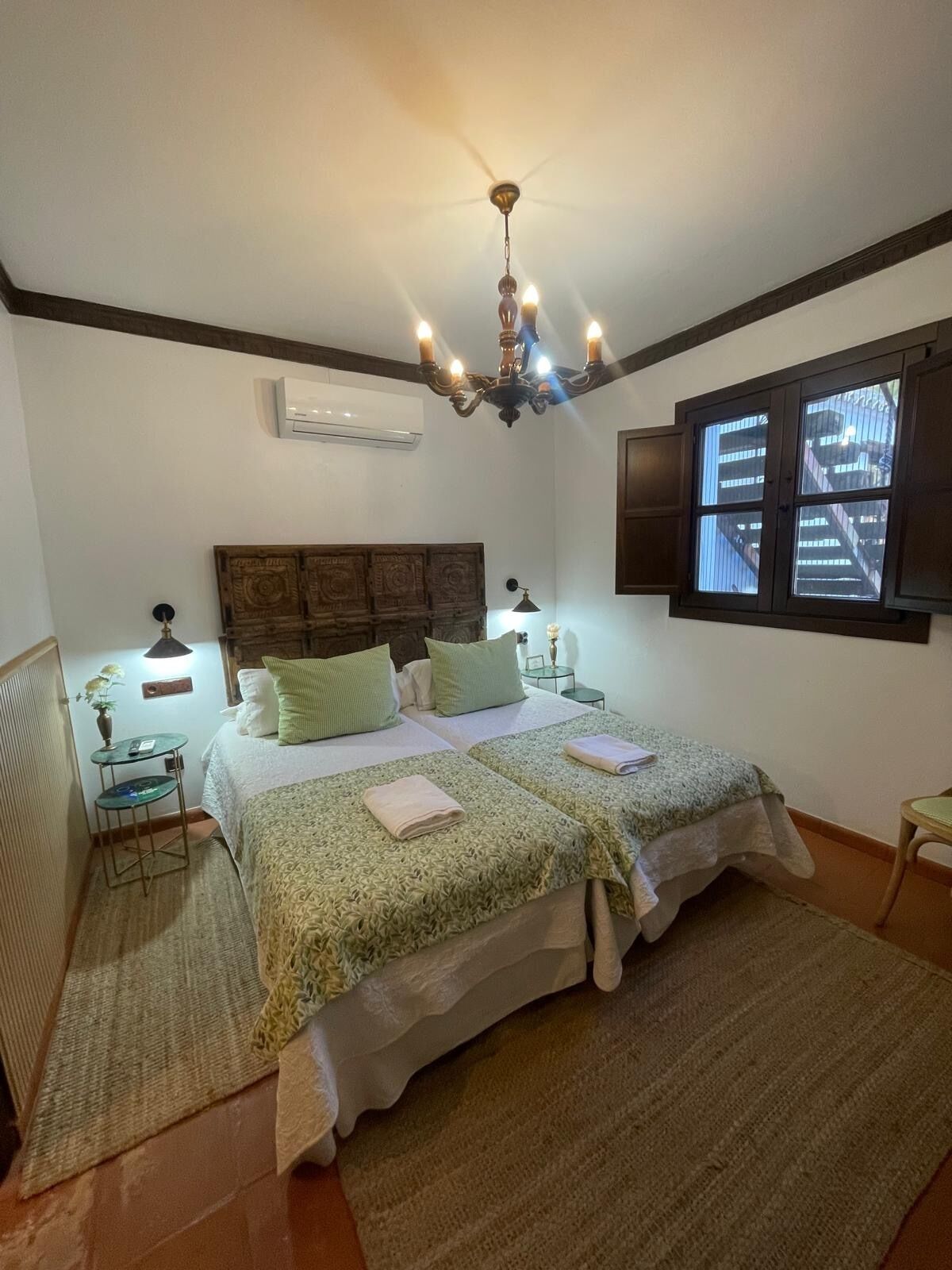 A bedroom with a large bed, a chandelier, a window with blinds, and a small table with a lamp.