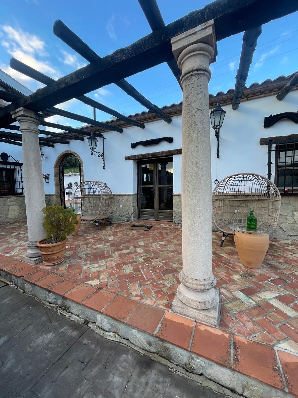A courtyard with a tiled floor, columns, and a pergola. There are potted plants and a wicker chair.