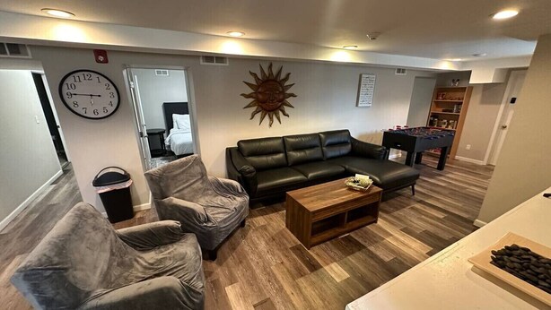 Un salon avec un canapé en cuir noir, deux fauteuils gris, une table basse en bois et une horloge murale.