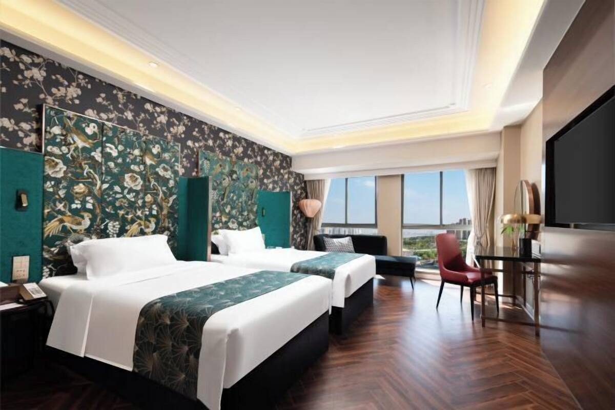 A hotel room with two beds, a desk, a chair, a TV, and a view of the city.