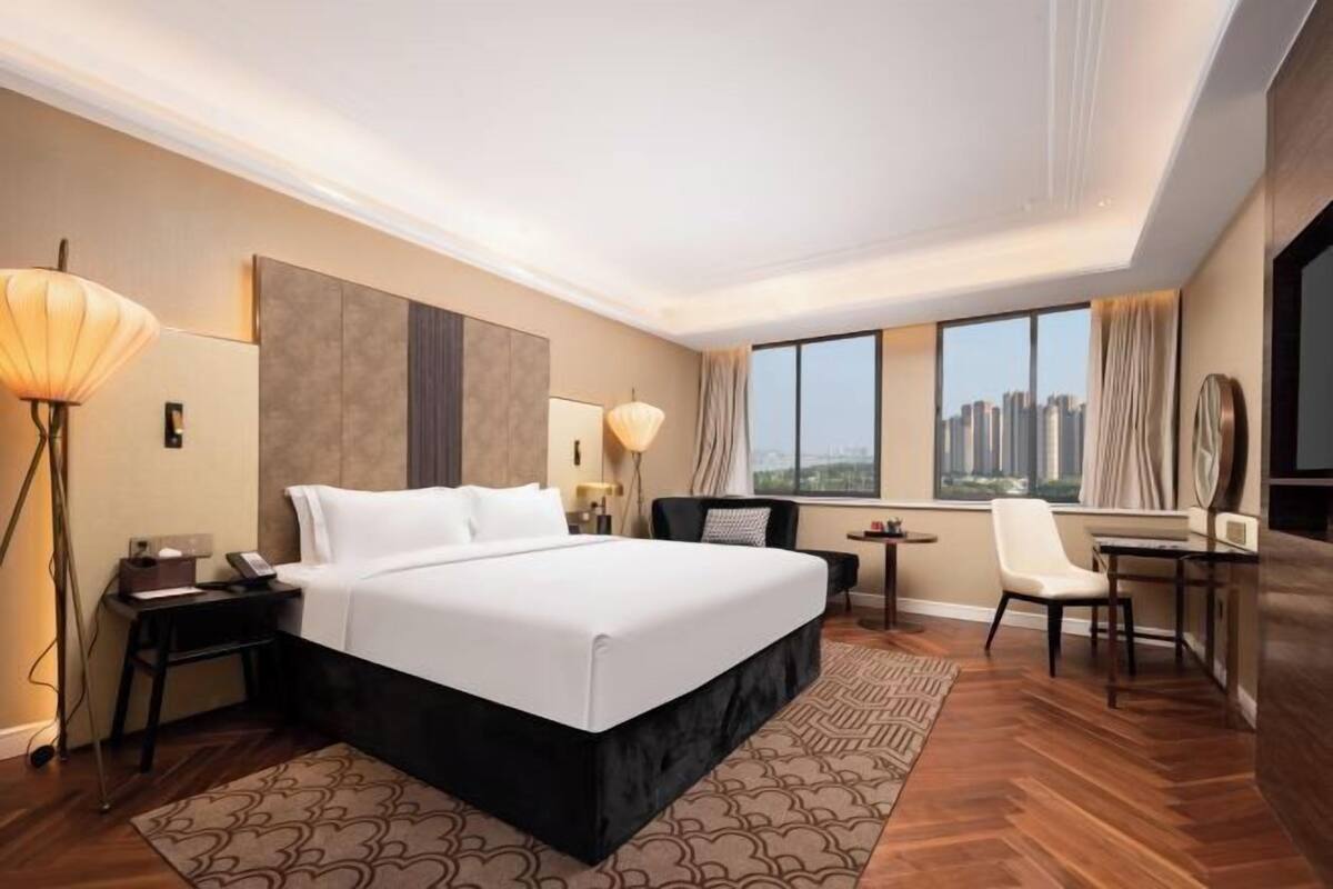 A hotel room with a large bed, a desk, a chair, a lamp, and a view of the city through large windows.