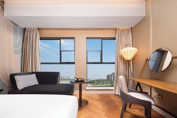 Habitación de hotel con cama, escritorio, una silla y vistas a la ciudad a través de amplios ventanales.