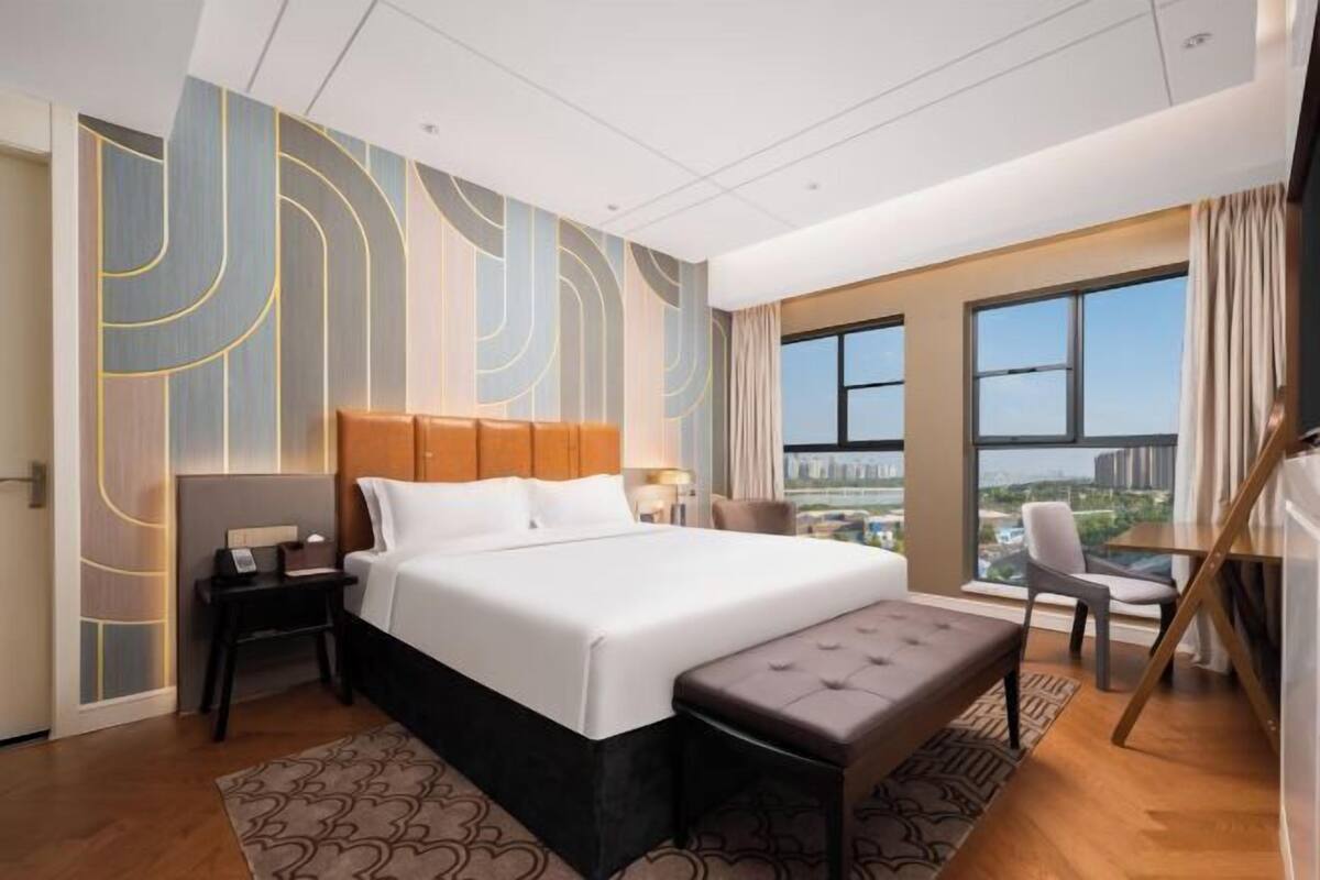 A modern hotel room with a large bed, a nightstand, a bench, a desk, and a view of the city.