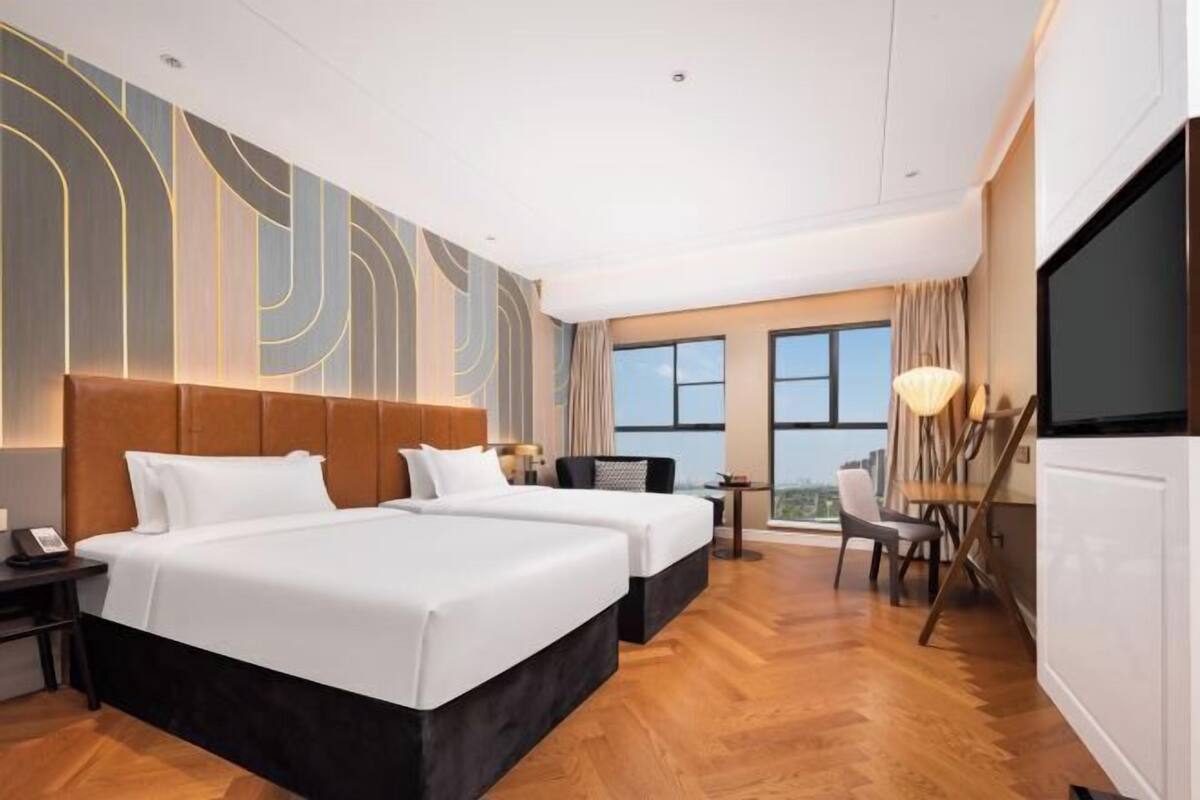 A hotel room with two beds, a desk, a chair, a TV, and a large window.