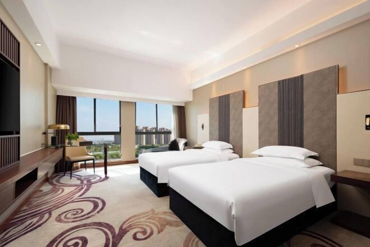 A hotel room with two beds, a desk, a chair, and a large window offering a city view.