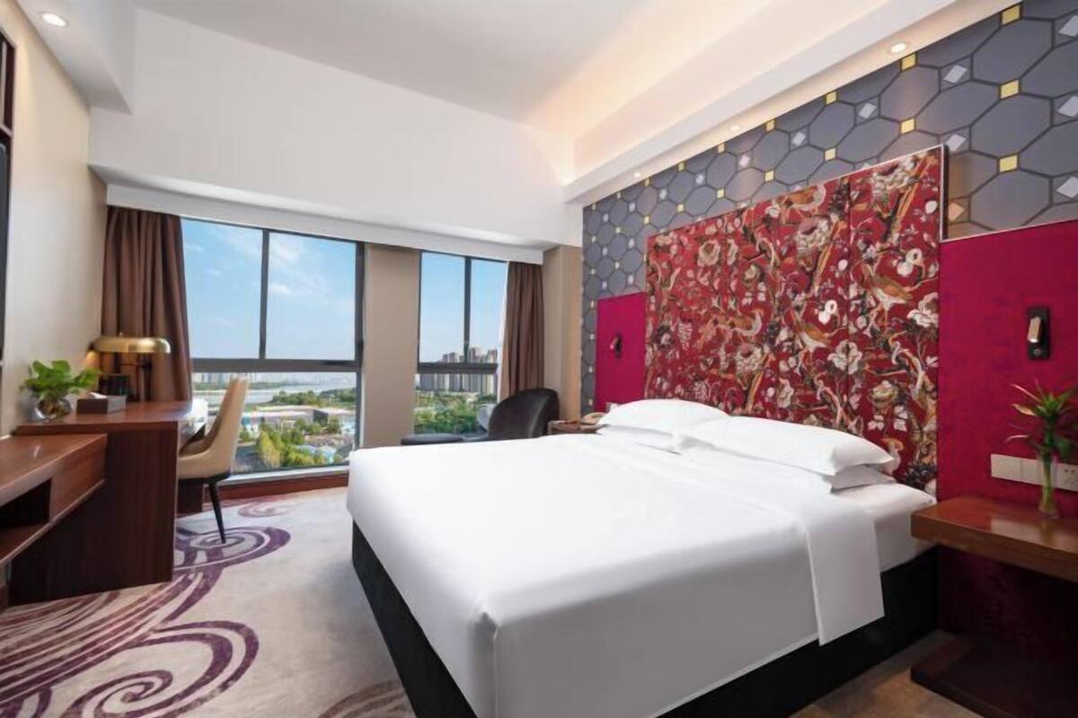 A hotel room with a large bed, a desk, and a chair. There is a window with a view of the city.