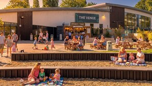 An outdoor seating area with tables and benches, a children's play area, and a building with a sign reading 'THE VENUE'. - Nature's Nook - Billing Aquadrome (Northampton)