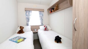 A compact room with two beds, a window with curtains, a shelf with toys, and a clock on the bedside table. - Nature's Nook - Billing Aquadrome (Northampton)