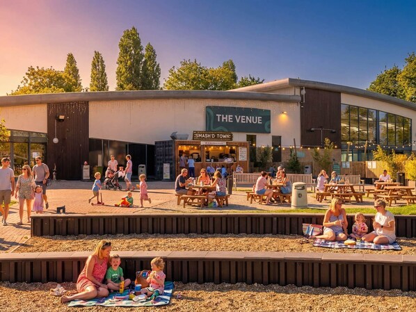 An outdoor seating area with tables and benches, a children's play area, and a building with a sign reading 'THE VENUE'. - Nature's Nook - Billing Aquadrome (Northampton)