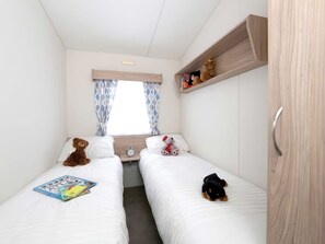 A compact room with two beds, a window with curtains, a shelf with toys, and a clock on the bedside table. - Nature's Nook - Billing Aquadrome (Northampton)