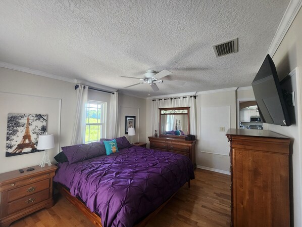 Un dormitorio con colcha morada, cómoda de madera y ventilador de techo.